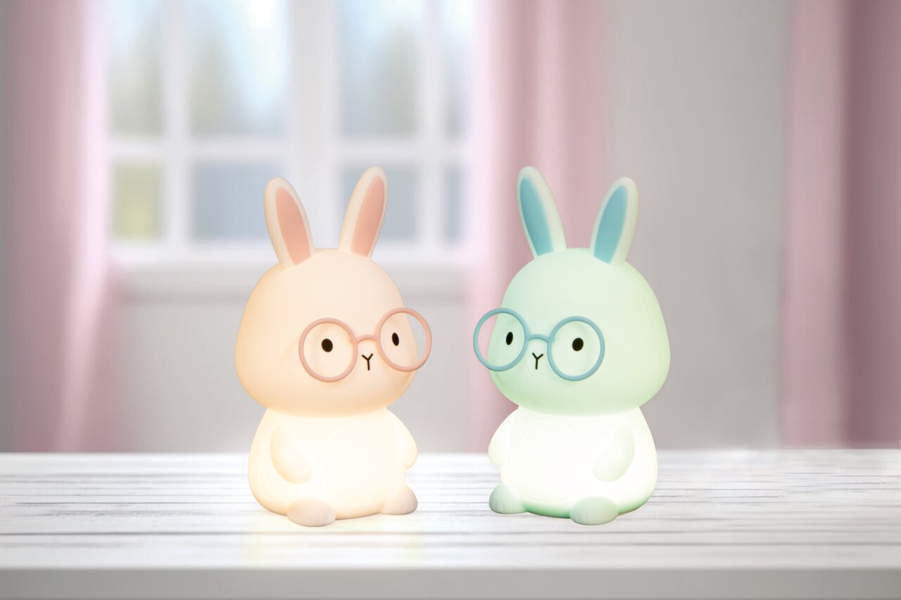 Zwei leuchtende Nachtlichter in Form von Hasen mit Brillen, auf einem Tisch vor einem Fenster, frontal betrachtet.