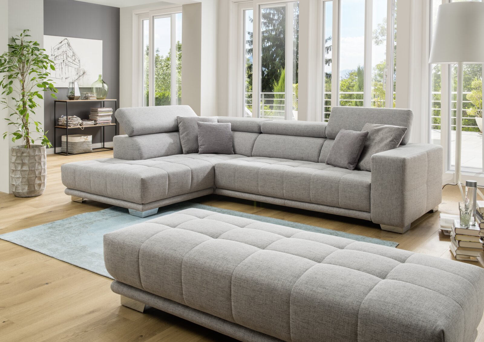 XORA Ecksofa CAORLE Moderne graue Polsterrundecke in einem hellen Wohnzimmer mit großen Fenstern, seitliche Perspektive