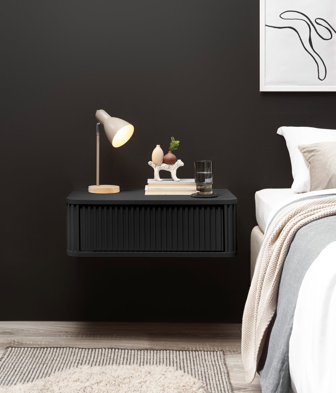 Z2 Nachtkommode GARI Schwebender, schwarzer Nachttisch mit geriffelter Front an dunkler Wand, dekoriert mit Lampe, Büchern und Glas neben einem Bett, Frontansicht.