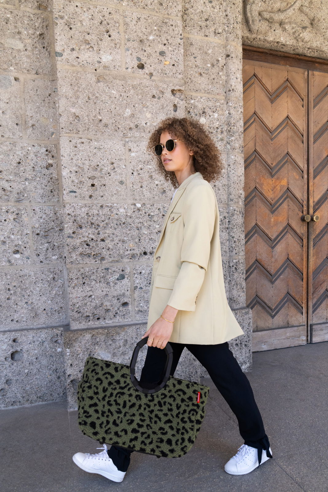 Person in heller Jacke und Sonnenbrille trägt eine große, grüne Tasche mit Leopardenmuster in der Hand, seitliche Perspektive.