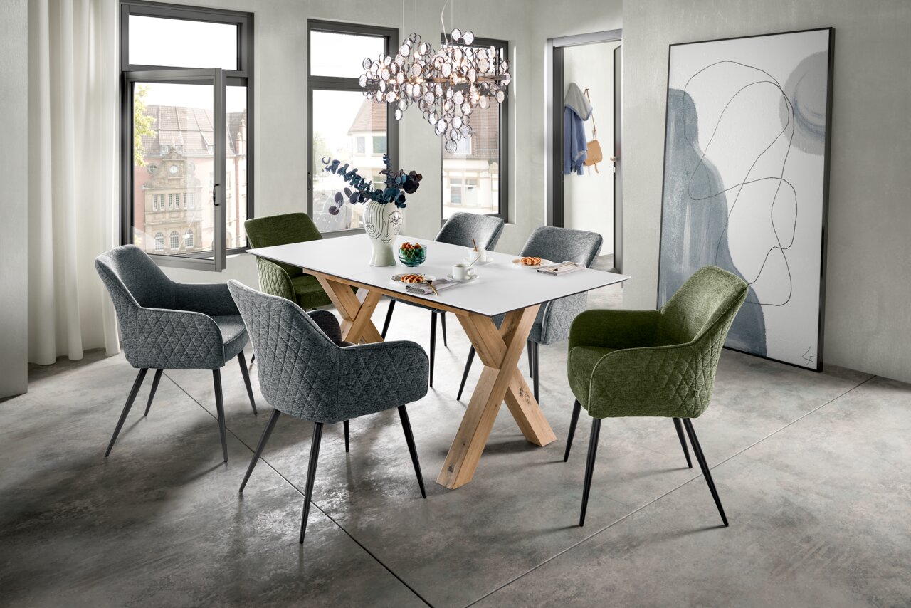 Moderner Esstisch mit weißer Tischplatte und Holzbeinen, umgeben von gepolsterten Stühlen in Grau und Grün, in einem hellen Raum mit großen Fenstern, aus seitlicher Perspektive.