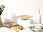 Keramik-Pastateller in Weiß mit zart apricotfarbenem Innenrand auf weißem Podest, seitliche Perspektive; im Vordergrund weitere Teller, Tomaten, Nudeln und eine weiße Ölflasche vor hellem Hintergrund.