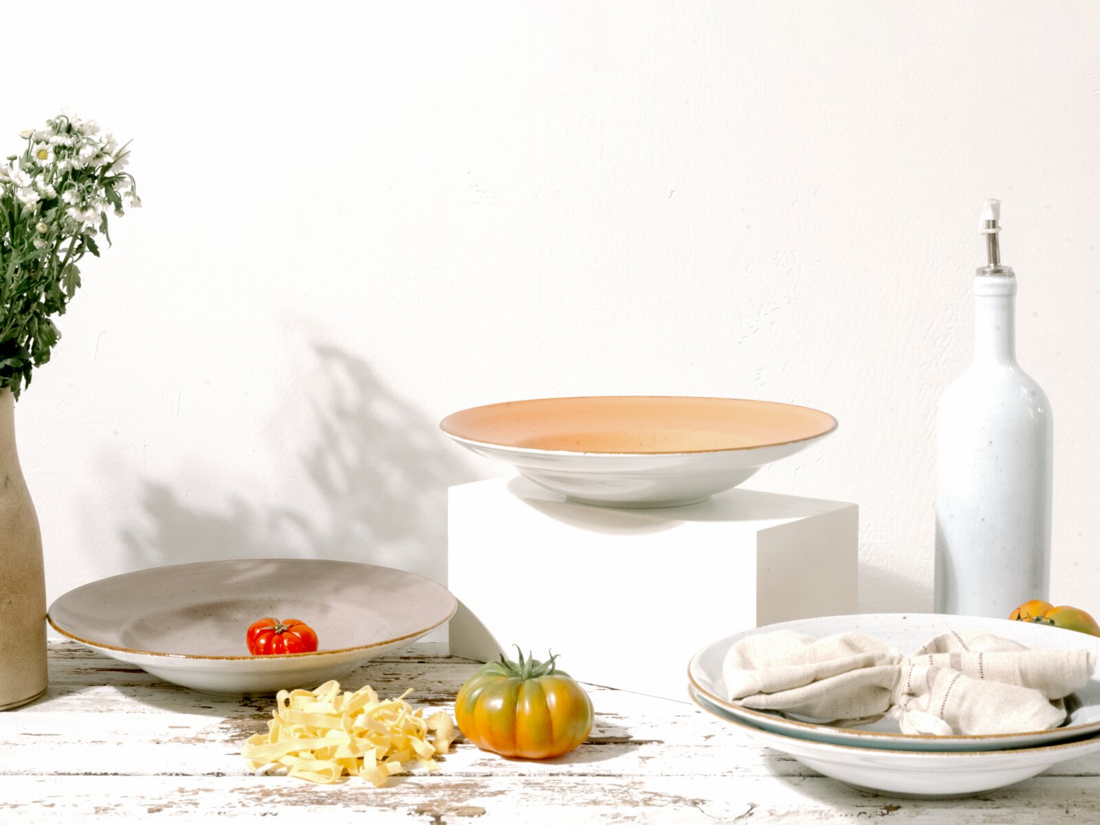 Creatable Pastateller 4-tlg VINTAGE NATURE Keramik-Pastateller in Weiß mit zart apricotfarbenem Innenrand auf weißem Podest, seitliche Perspektive; im Vordergrund weitere Teller, Tomaten, Nudeln und eine weiße Ölflasche vor hellem Hintergrund.