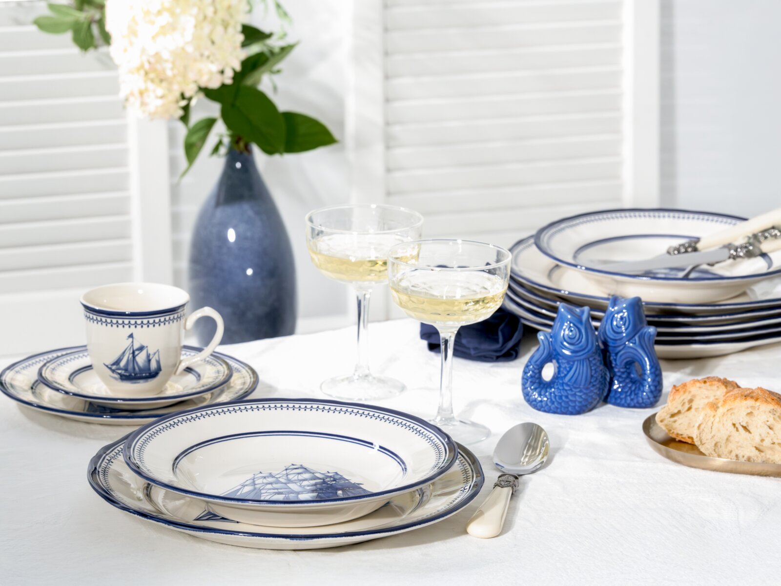 Schlichtes blau-weißes Geschirrset mit maritimen Schiffmotiven auf gedecktem Tisch, inklusive Tasse, Teller, Schalen, Sektgläsern und blauen Fisch-Salzstreuern; Perspektive: leicht erhöhte Frontansicht auf Tischhöhe.