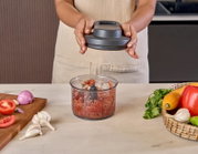 Persoon in schort houdt het deksel van een keukenhakmolen boven een transparante kom met fijngesneden tomaten, ui en kruiden op het aanrecht; frontaal aanzicht op werkhoogte.
