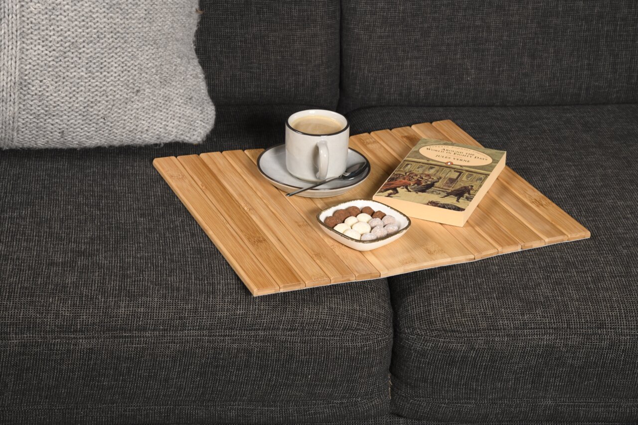 Bambus Sofaablage auf einem grauen Sofa mit einer Tasse Kaffee, einem Buch und einer Schale mit Süßigkeiten, aus der Vogelperspektive.