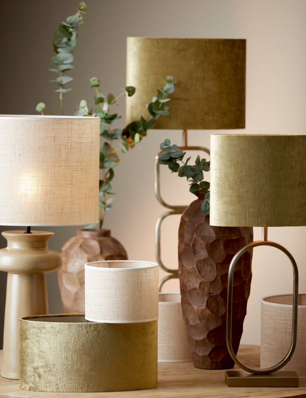Mehrere Lampenschirme in verschiedenen Größen und Farben, darunter ein zylindrischer Lampenschirm in Beige und Gold, auf einem Tisch arrangiert. Die Perspektive zeigt die Lampenschirme von vorne.