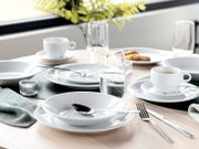 Modern gedeckter Esstisch mit weißem Porzellan-Geschirr (Teller, Schalen, Tassen), Gläsern und Besteck auf hellem Holztisch, schräg von oben aus der Tischperspektive fotografiert.