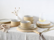 Modernes Keramik-Geschirrset in Beige und Weiß mit Tellern und Schalen auf Holzbrett, daneben Weinglas und goldenes Besteck auf hellem Tisch, fotografiert in leicht erhöhter Frontansicht.