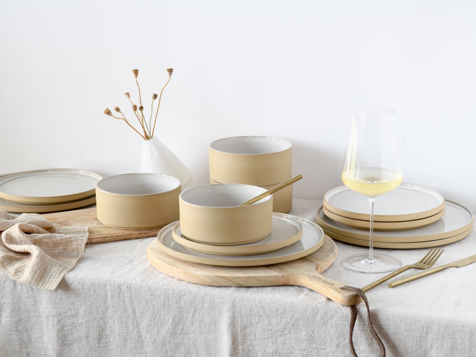 Modernes Keramik-Geschirrset in Beige und Weiß mit Tellern und Schalen auf Holzbrett, daneben Weinglas und goldenes Besteck auf hellem Tisch, fotografiert in leicht erhöhter Frontansicht.