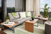Moderne Gartenlounge mit grauem Sofa, grünen und weißen Kissen, Holztischen und Pflanzen auf einem Balkon mit Aussicht.