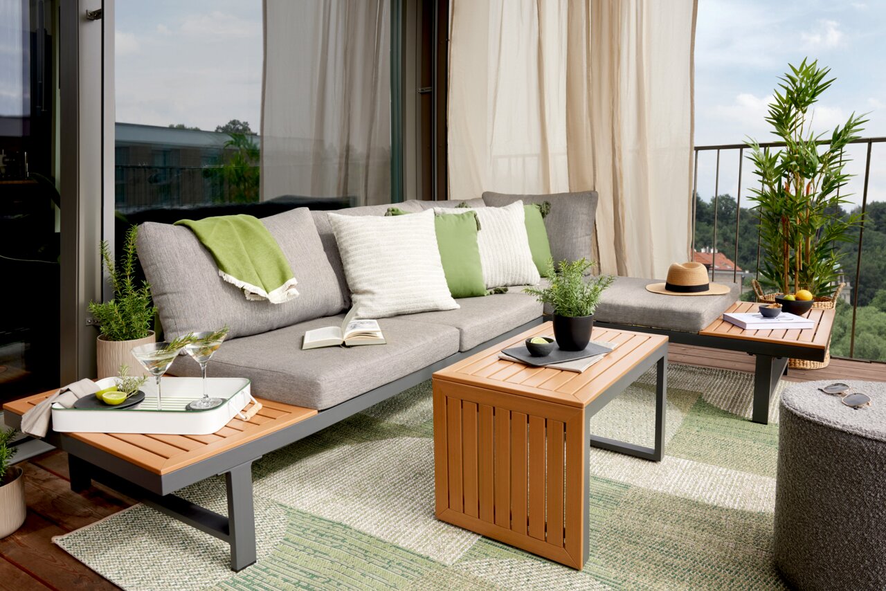 Moderne Gartenlounge mit grauem Sofa, grünen und weißen Kissen, Holztischen und Pflanzen auf einem Balkon mit Aussicht.