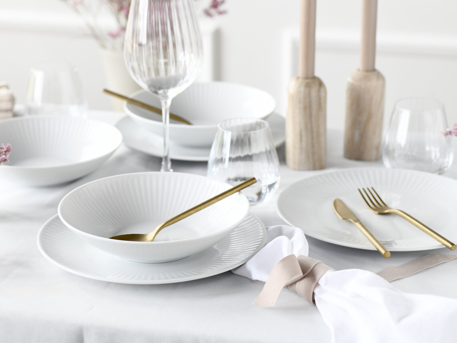 Elegant gedeckter Tisch mit weißen geriffelten Tellern und Schalen, goldfarbenem Besteck, Weinglas und Wasserglas sowie hellen Holz-Kerzenhaltern; Perspektive: leicht schräge Ansicht von oben.
