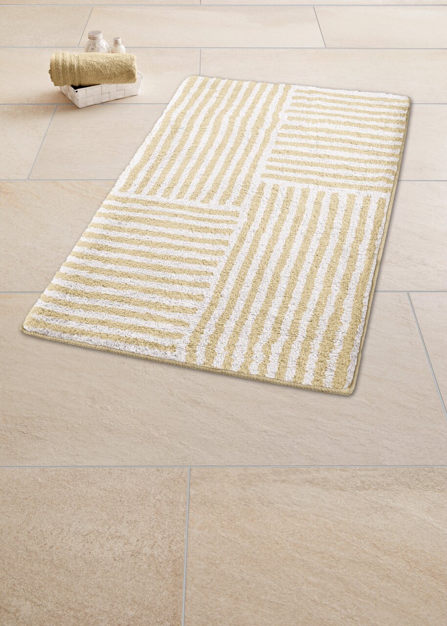 Beige-weißer Badteppich mit Streifenmuster auf Fliesenboden, perspektivisch von oben fotografiert.