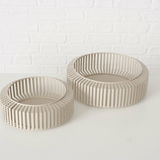 Zwei moderne, runde Schalen aus beigem Material mit einem Gitterdesign, auf einer weißen Oberfläche vor einer weißen Ziegelwand, aus einer leicht erhöhten Perspektive fotografiert.