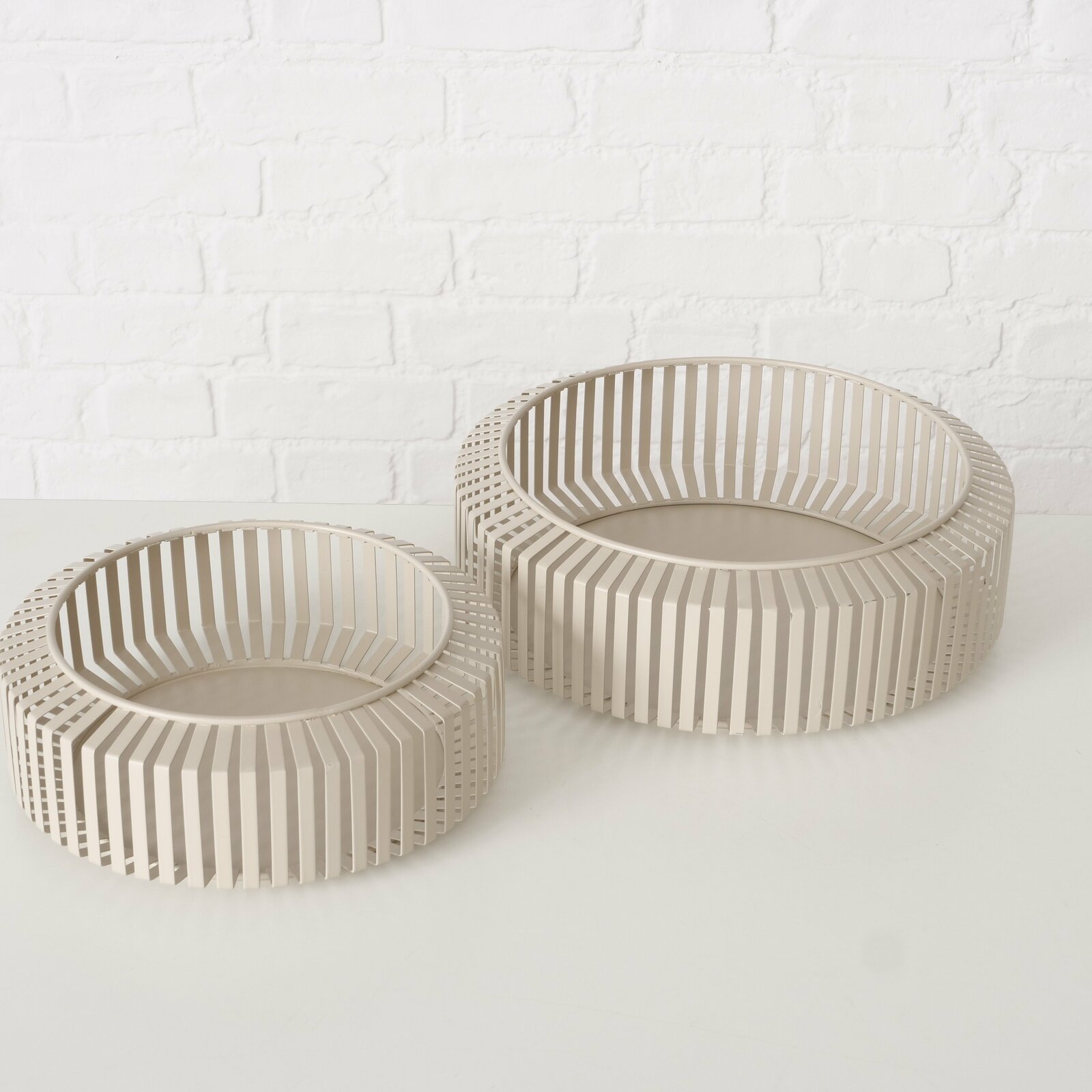 Zwei moderne, runde Schalen aus beigem Material mit einem Gitterdesign, auf einer weißen Oberfläche vor einer weißen Ziegelwand, aus einer leicht erhöhten Perspektive fotografiert.