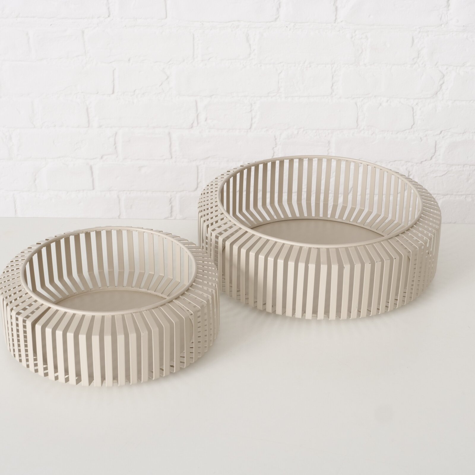 Zwei moderne, runde Schalen aus beigem Material mit einem Gitterdesign, auf einer weißen Oberfläche vor einer weißen Ziegelwand, aus einer leicht erhöhten Perspektive fotografiert.