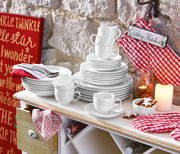 Ein stilvolles weißes Kombiservice-Set, bestehend aus Tellern, Tassen und Schalen, auf einem Tisch in einer gemütlichen Landhausküche. Perspektive von vorne.