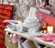 Ein stilvolles weißes Kombiservice-Set, bestehend aus Tellern, Tassen und Schalen, auf einem Tisch in einer gemütlichen Landhausküche. Perspektive von vorne.