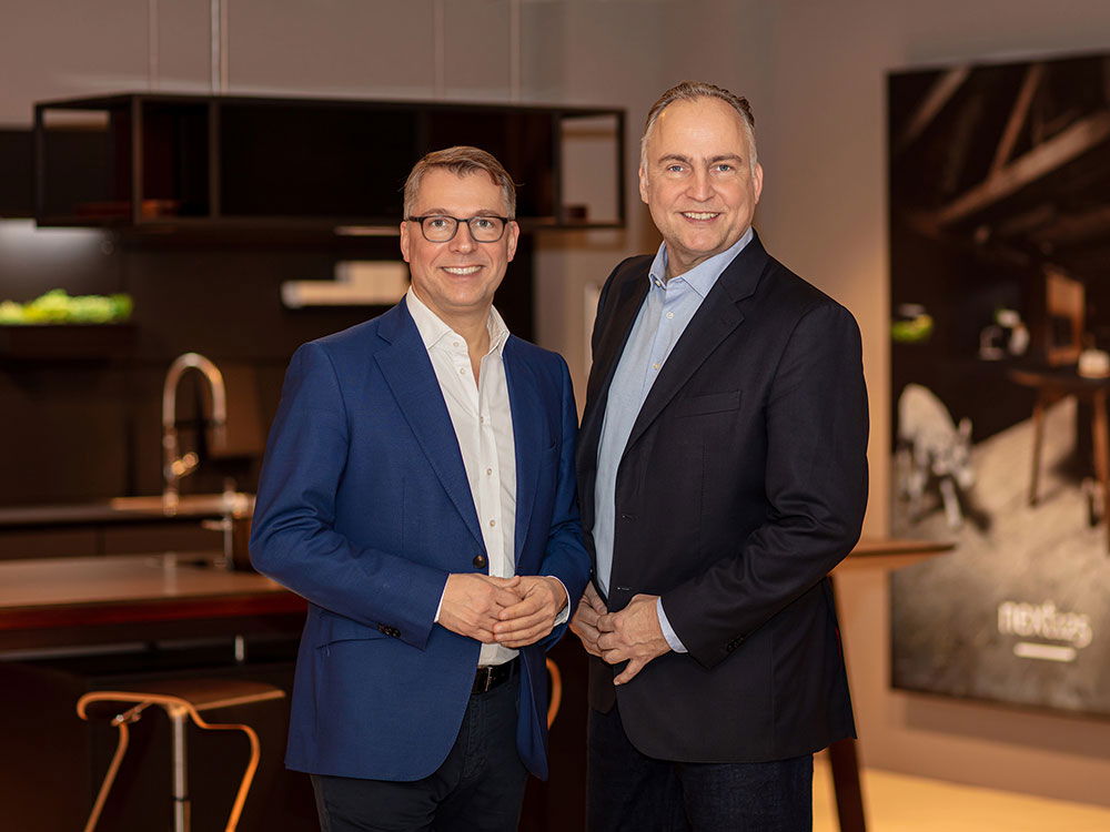 Zwei Männer in Anzügen lächeln in einer modernen Küchenumgebung in die Kamera.