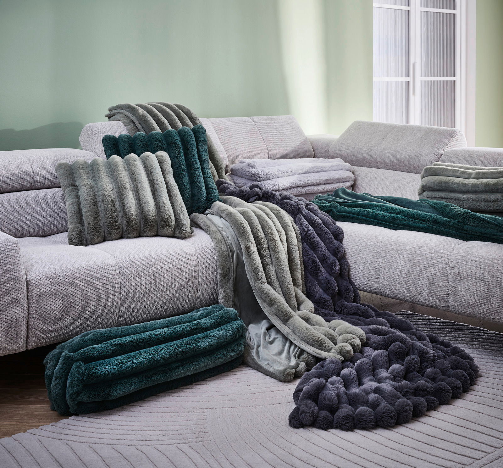 Luxuriöse Felldecken und Kissen in verschiedenen Grüntönen und Grau auf einem hellen Sofa, seitliche Perspektive.