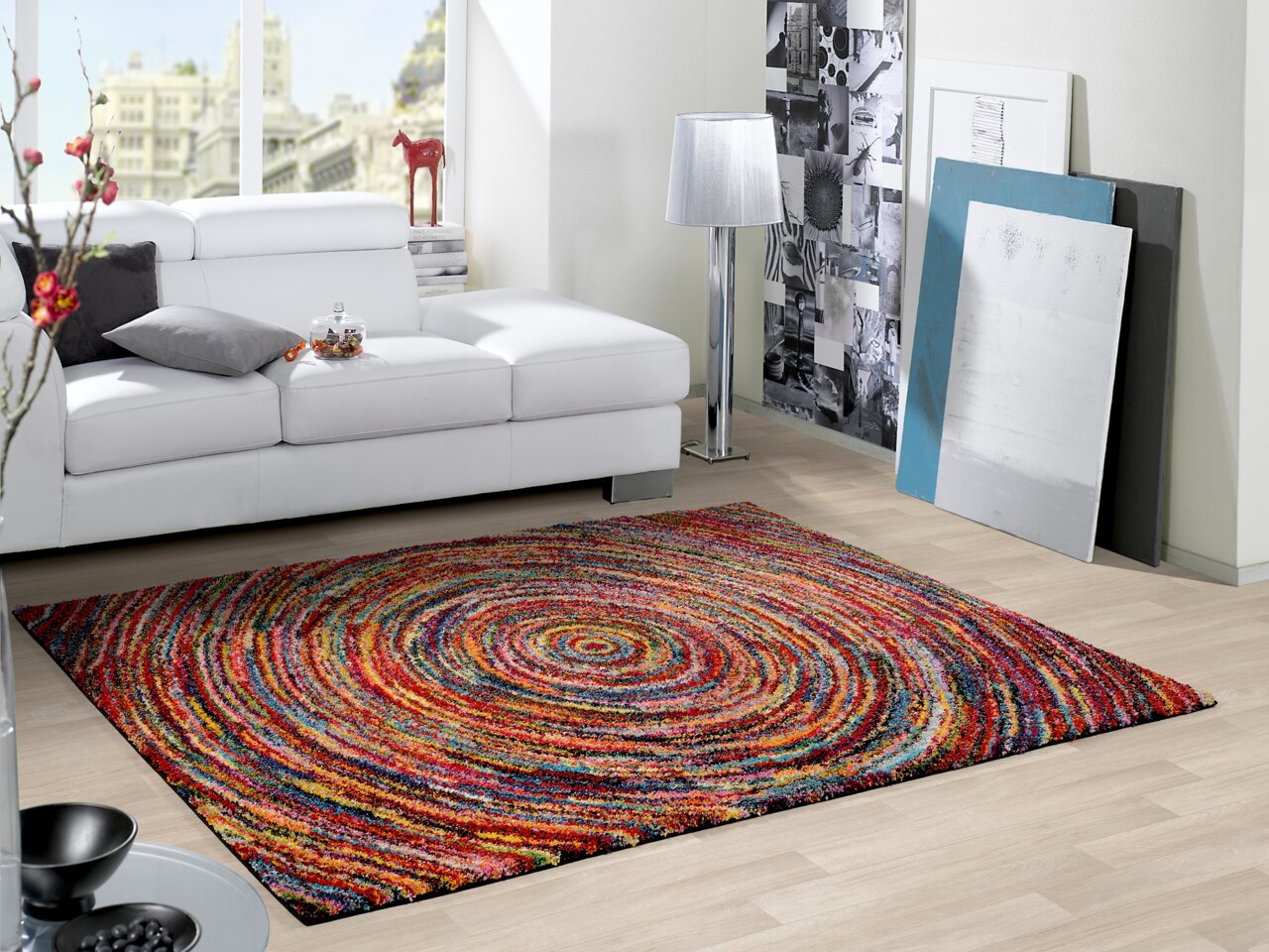 Bunter, runder Teppich mit spiralförmigem Muster in einem modernen Wohnzimmer mit weißem Sofa und dekorativen Elementen, aus der Perspektive von schräg oben.