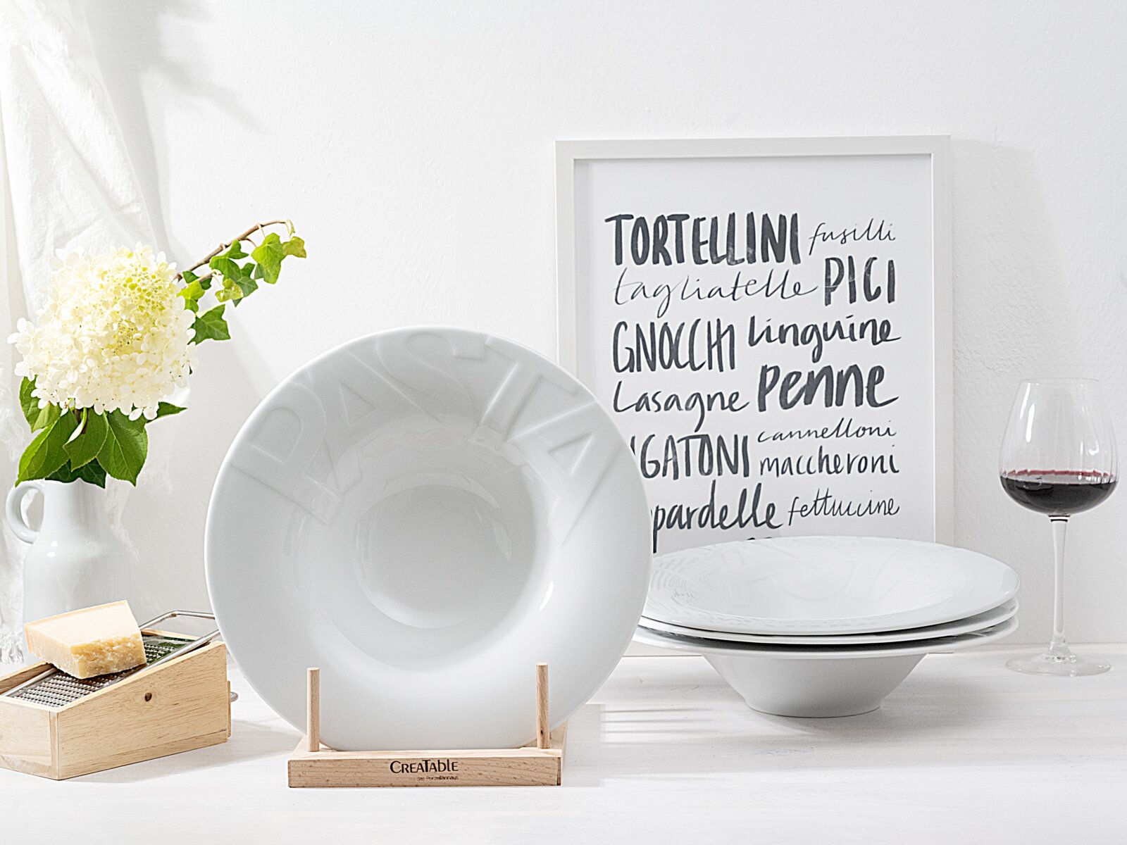 Creatable Pastateller 4-tlg GOURMET Weißer tiefer Pastateller aus Porzellan mit dezentem Relief-Schriftzug, frontal in einem Holzständer präsentiert; im Hintergrund weitere weiße Teller, ein Pasta-Poster und ein Weinglas (Perspektive: Frontalansicht).