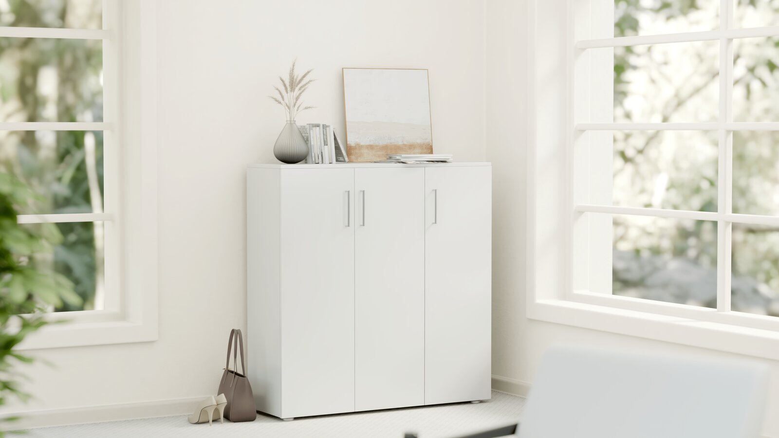 Moderano Schuhschrank breit SHOES Weißer Schuhschrank mit drei Türen in einer Ecke eines hellen Raumes, dekoriert mit einer Vase, Büchern und einem Bild auf der Oberseite, seitliche Perspektive