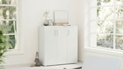 Weißer Schuhschrank mit drei Türen in einer Ecke eines hellen Raumes, dekoriert mit einer Vase, Büchern und einem Bild auf der Oberseite, seitliche Perspektive