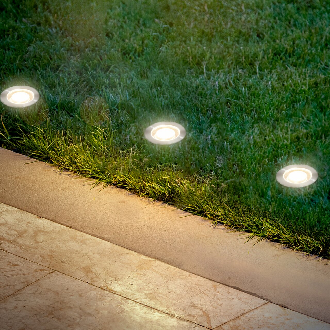 Bodenleuchten aus Edelstahl, eingebaut in eine Rasenfläche neben einem gepflasterten Weg, aus der Vogelperspektive betrachtet.