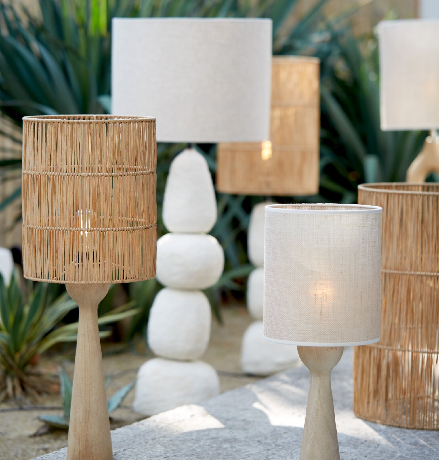 Verschillende lampen van natuurlijke materialen, waaronder een houten lampvoet met een ronde, geweven lampenkap, buiten opgesteld. Aanzicht van voren.