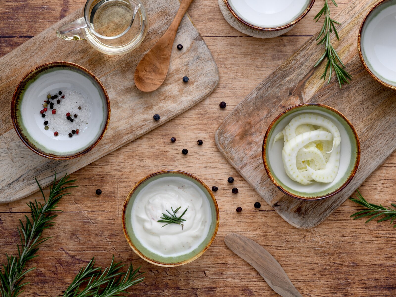 Draufsicht auf rustikales Food-Arrangement: Schalen mit Kräutercreme, grobem Salz mit Pfefferkörnern und Fenchelstreifen auf Holzbrettchen, dazu Ölkanne, Holzlöffel und Rosmarinzweige auf Holztisch