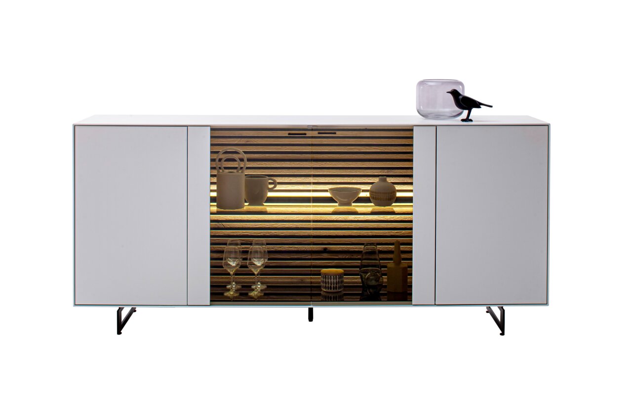 Modern dressoir met witte afwerking, glazen inzetstuk in het midden en decoratieve verlichting, gezien vanaf de voorkant.