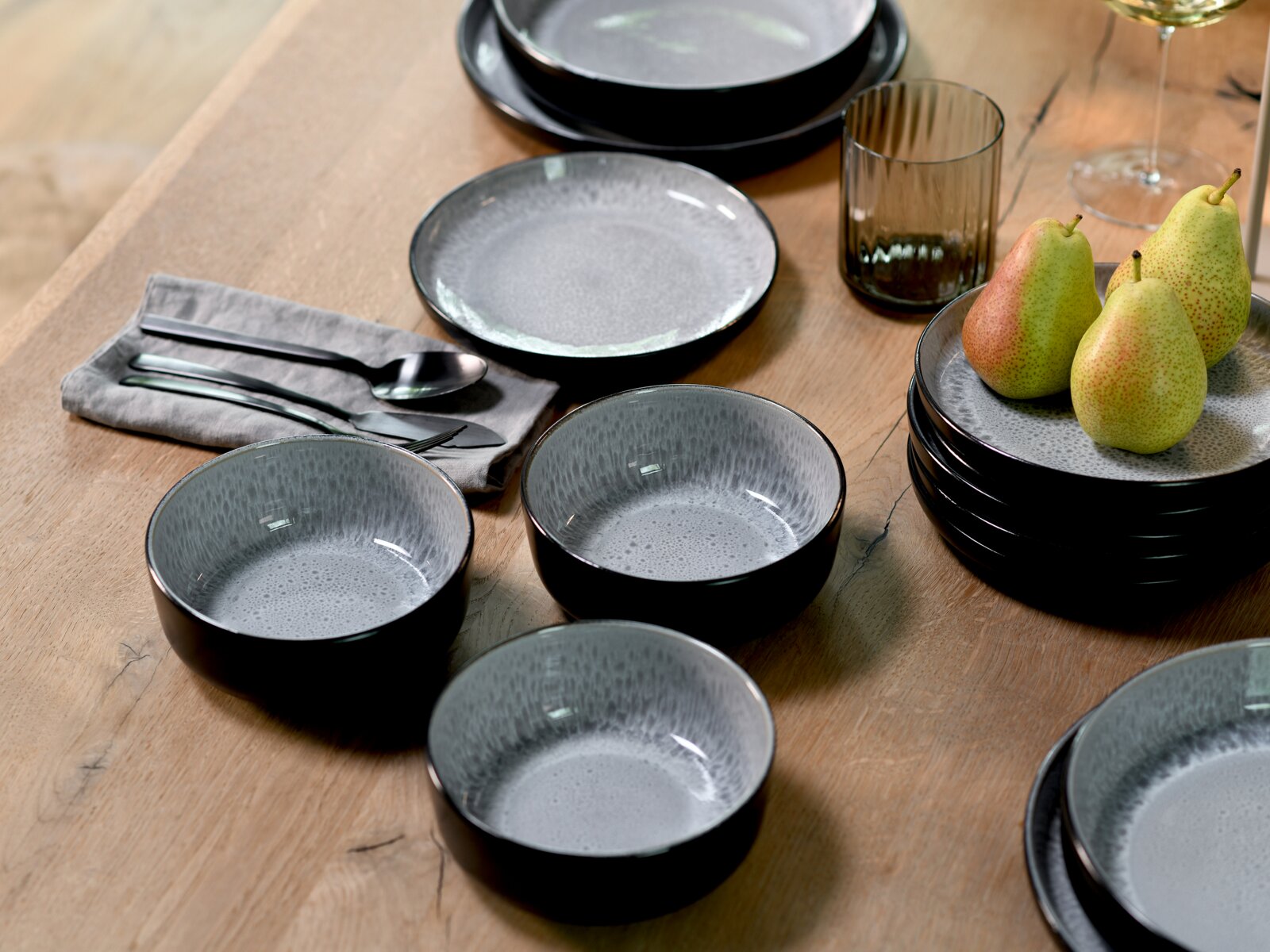 Schwarz-graues Keramik-Geschirrset mit Tellern und Schalen auf Holztisch gedeckt, mit Besteck, Glas und Birnen, aus leicht schräger Perspektive von oben