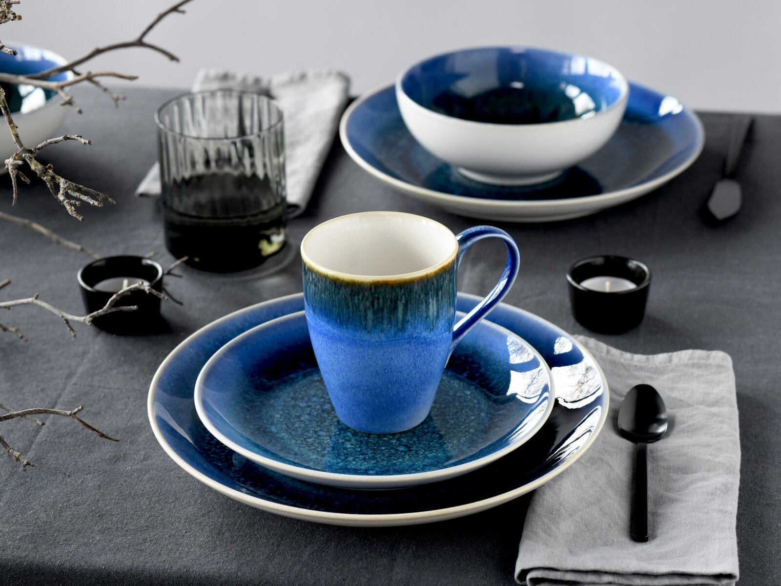 Blau glasiertes Keramik-Geschirrset mit Tasse auf Teller und großem Speiseteller sowie Schale im Hintergrund, gedeckter Tisch mit Glas, Löffel und Teelichtern, Perspektive: leicht von oben in schräger Frontansicht