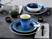 Blau glasiertes Keramik-Geschirrset mit Tasse auf Teller und großem Speiseteller sowie Schale im Hintergrund, gedeckter Tisch mit Glas, Löffel und Teelichtern, Perspektive: leicht von oben in schräger Frontansicht