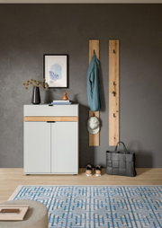 Modernes Flur-Set mit weißem Schuhschrank mit Holz-Akzent, zwei vertikalen Garderobenpaneelen aus Holz mit Haken, aufgehängter Jacke und Cap sowie Tasche und Schuhen auf grauer Wand, Frontansicht.