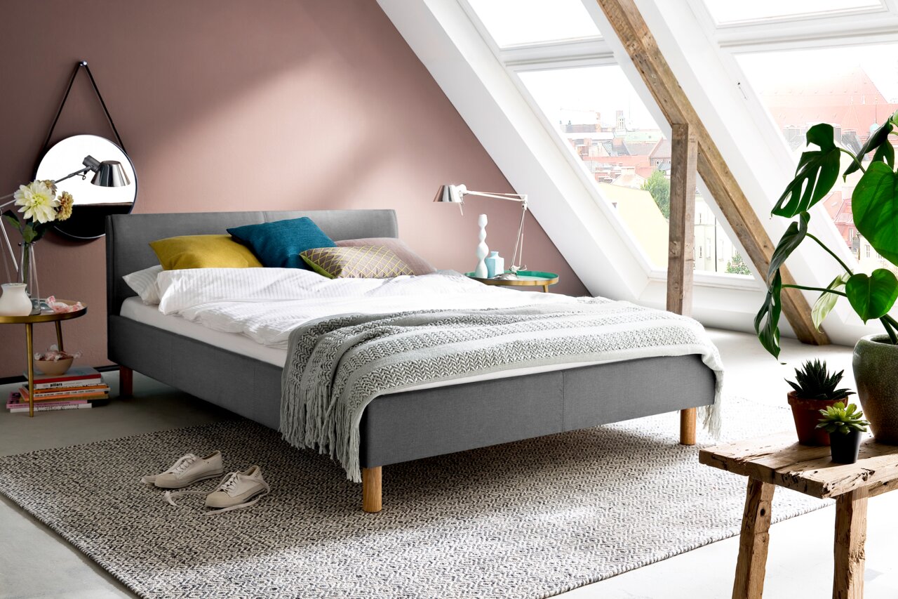 Graues Polsterbett in einem modernen Schlafzimmer mit Dachschräge, dekoriert mit bunten Kissen und einer Tagesdecke, seitliche Perspektive.