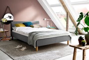 Graues Polsterbett in einem modernen Schlafzimmer mit Dachschräge, dekoriert mit bunten Kissen und einer Tagesdecke, seitliche Perspektive.