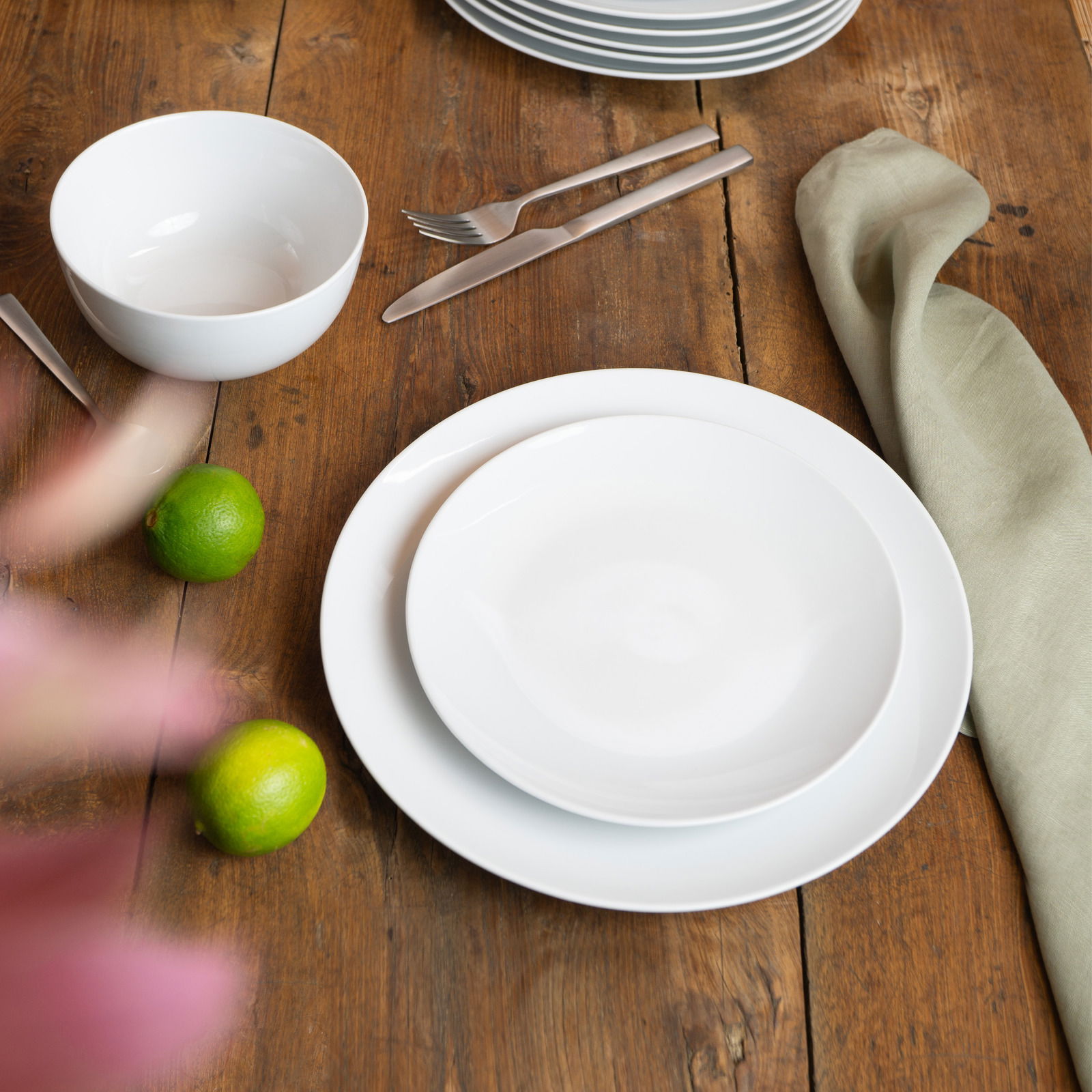 Weißes Geschirrset auf einem Holztisch, bestehend aus einem flachen Teller, einem tiefen Teller und einer Schüssel, mit Besteck und Limetten als Dekoration, aus der Vogelperspektive fotografiert.