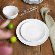 Weißes Geschirrset auf einem Holztisch, bestehend aus einem flachen Teller, einem tiefen Teller und einer Schüssel, mit Besteck und Limetten als Dekoration, aus der Vogelperspektive fotografiert.