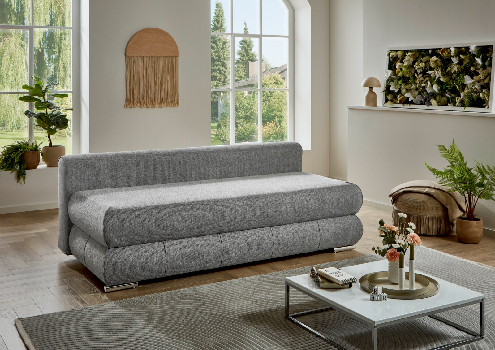 Modernes graues Schlafsofa/Polstersofa mit abgerundeten Kanten im hellen Wohnzimmer, Perspektive: Frontansicht leicht schräg von links.