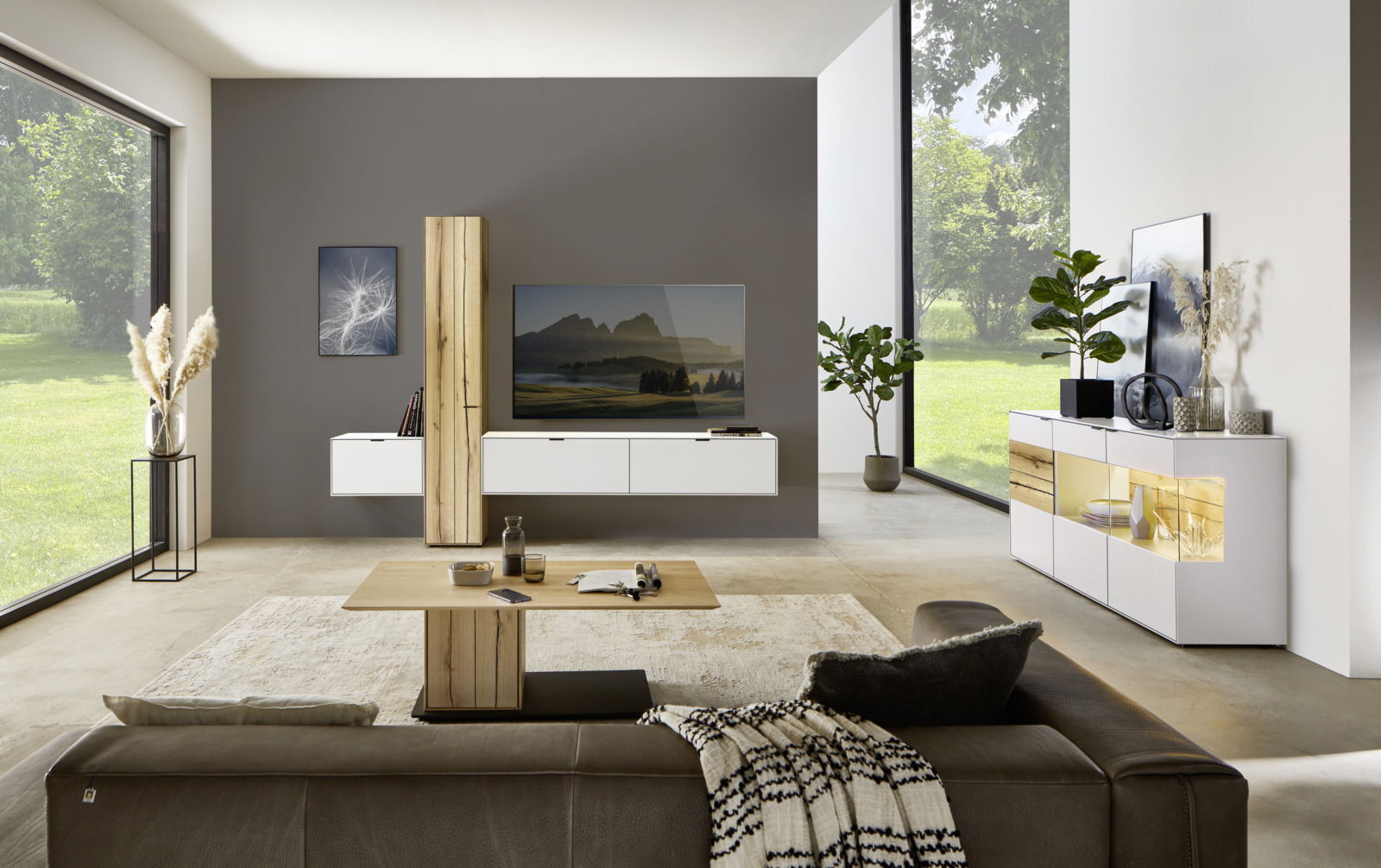 Moderne Wohnzimmeransicht mit der Dieter Knoll Wandkombination ISEO in Weiß und Holzoptik, frontal betrachtet. Die Kombination besteht aus hängenden Schränken und einem TV-Element, umgeben von stilvollen Dekorationen und großen Fenstern mit Blick ins Grün