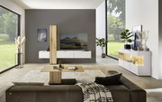 Moderne Wohnzimmeransicht mit der Dieter Knoll Wandkombination ISEO in Weiß und Holzoptik, frontal betrachtet. Die Kombination besteht aus hängenden Schränken und einem TV-Element, umgeben von stilvollen Dekorationen und großen Fenstern mit Blick ins Grün