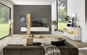 Moderne Wohnzimmeransicht mit der Dieter Knoll Wandkombination ISEO in Weiß und Holzoptik, frontal betrachtet. Die Kombination besteht aus hängenden Schränken und einem TV-Element, umgeben von stilvollen Dekorationen und großen Fenstern mit Blick ins Grün