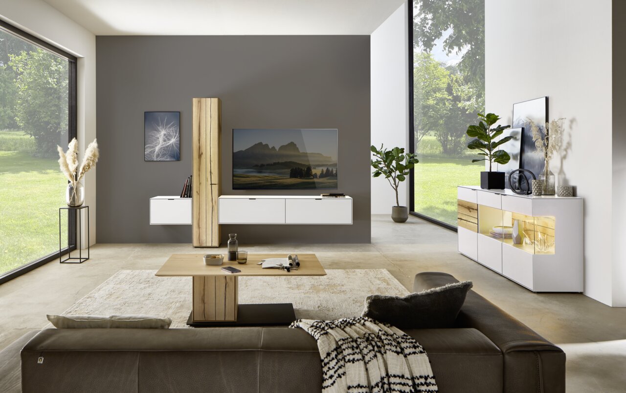 Moderne Wohnzimmeransicht mit der Dieter Knoll Wandkombination ISEO in Weiß und Holzoptik, frontal betrachtet. Die Kombination besteht aus hängenden Schränken und einem TV-Element, umgeben von stilvollen Dekorationen und großen Fenstern mit Blick ins Grün