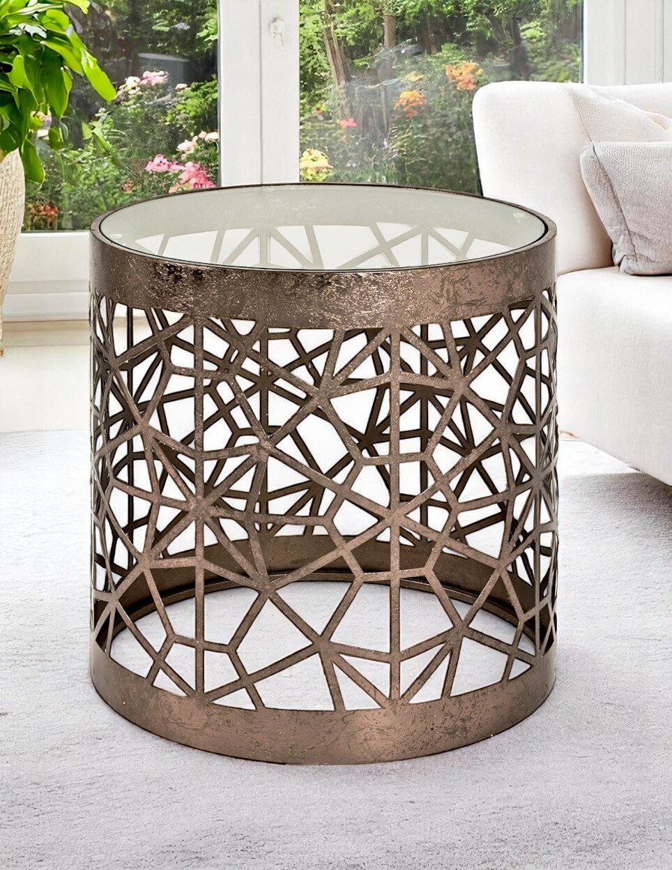 Runder Beistelltisch/Couchtisch aus bronzefarbenem Metall mit geometrischem Cut-out-Gitter und Glasplatte, in schräger Frontansicht im Wohnzimmer neben einem hellen Sofa.