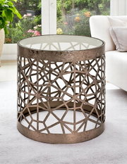 Runder Beistelltisch/Couchtisch aus bronzefarbenem Metall mit geometrischem Cut-out-Gitter und Glasplatte, in schräger Frontansicht im Wohnzimmer neben einem hellen Sofa.