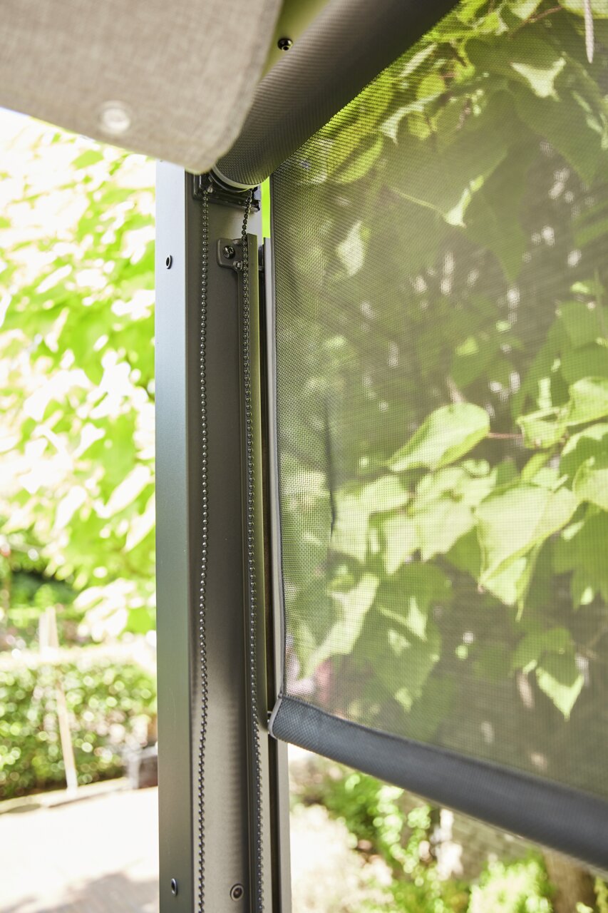 Detailfoto van een rolgordijn met kettingmechanisme, op de achtergrond groene bladeren.