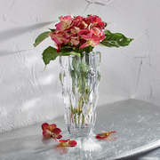 Kristallvase mit facettierter Oberfläche, gefüllt mit rosa Blumen, auf einem Tisch vor einer strukturierten Wand, seitliche Perspektive